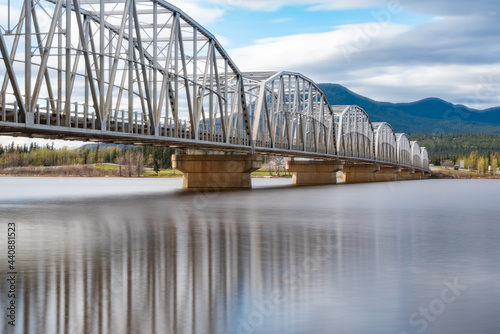Large man made structure steel bridge spanning across Nisutlin Bay in township of Teslin flowing to the Yukon River in northern Canada during spring summer time with cloudy mountains background. © Scalia Media