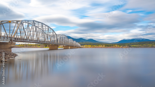 Large man made structure steel bridge spanning across Nisutlin Bay in township of Teslin flowing to the Yukon River in northern Canada during spring summer time with cloudy mountains background. photo