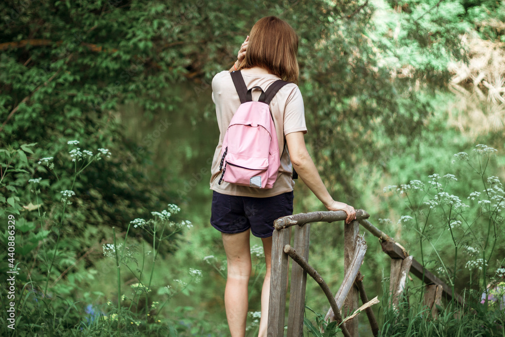 A young woman with short hair and a pink backpack stands with her back to the camera on a small wooden bridge near a river in the woods. Selective Focus