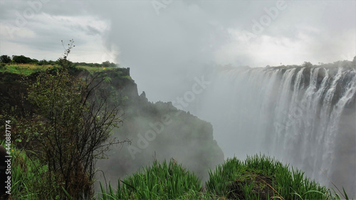 Powerful streams of water fall into the gorge. A dense fog stands over the abyss. Tiny silhouettes of people are visible on the plateau. Lush green grass in the foreground. Victoria Falls. Zambia