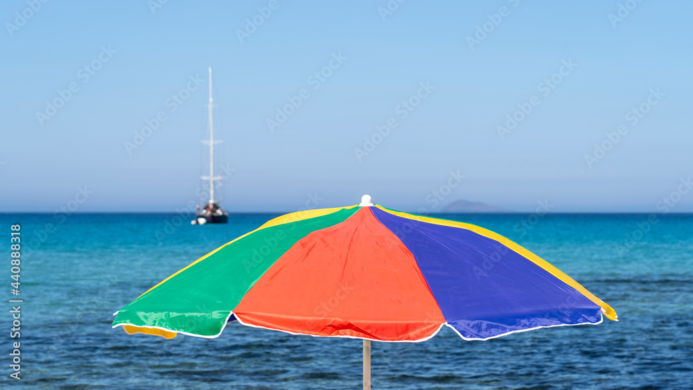 Isolated green, red, blue and yellow beach umbrella. Blue sky. Relaxing context. Summer holidays at the sea. General contest and location. Turquoise sea in the background. Multicolor umbrella