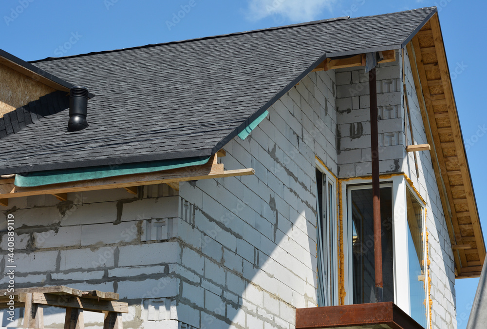 A brick house under construction with a roof covered with asphalt shingles, roof ventilation cap installed, and unfinished soffit.