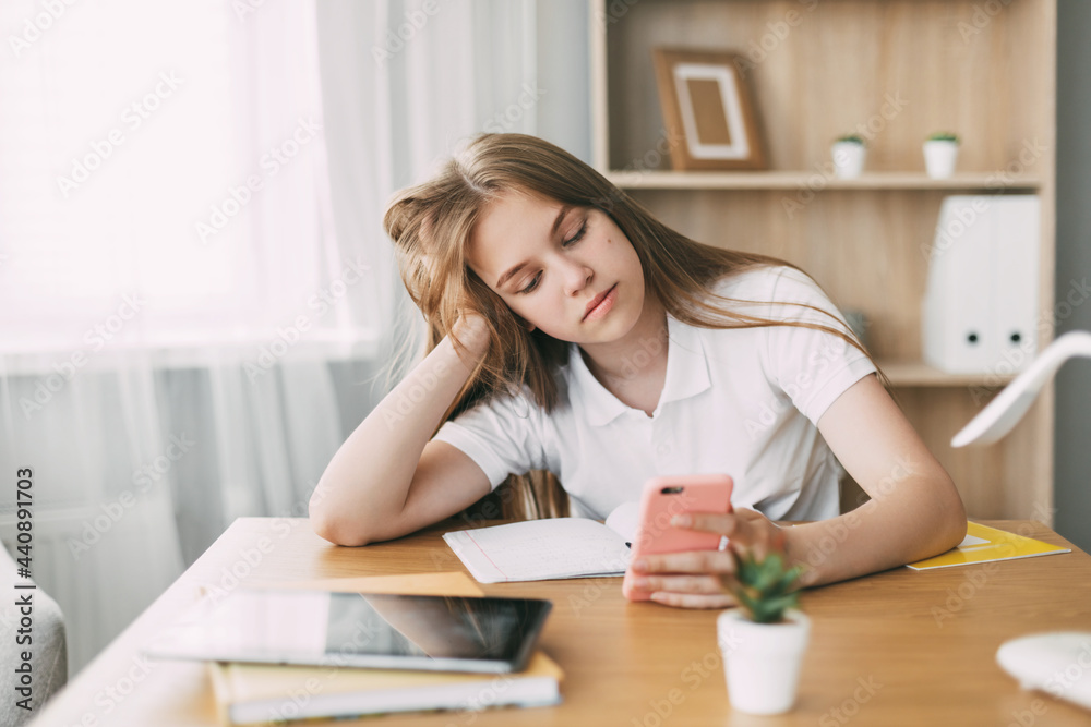 Cute schoolgirl is tired of doing homework and is looking for a solution to the problem online using the phone Home education, home training Cute schoolgirl is tired of doing homework and is looking for a solution to the problem online using the phone Home education, home training
