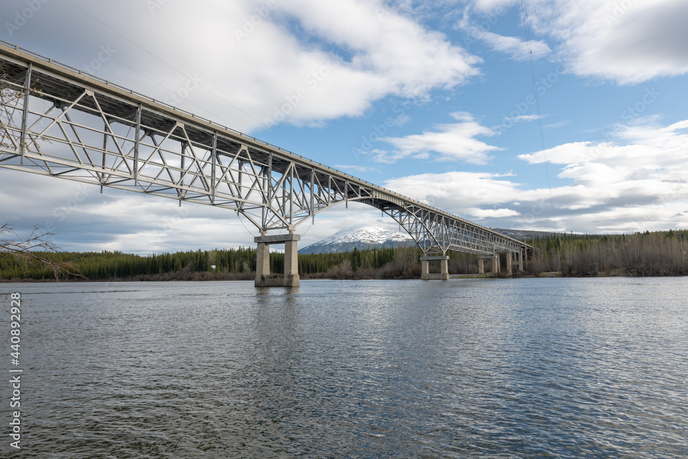 Johnsons Crossing, Teslin River steel Bridge on the Alaska Highway during spring summer time with cloudy, blue sky day and magnificent, huge structure over the flowing water below. 