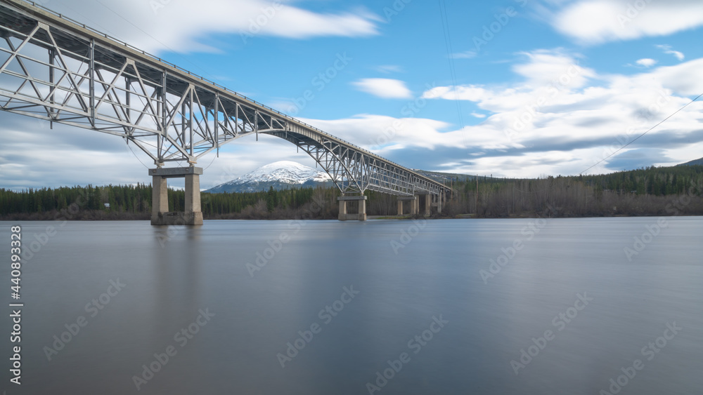 Johnsons Crossing, Teslin River steel Bridge on the Alaska Highway during spring summer time with cloudy, blue sky day and magnificent, huge structure over the flowing water below. 