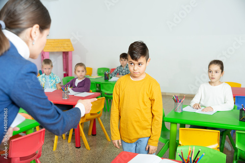Upset boy listening reprimanding of strict female teacher during lesson in primary school photo