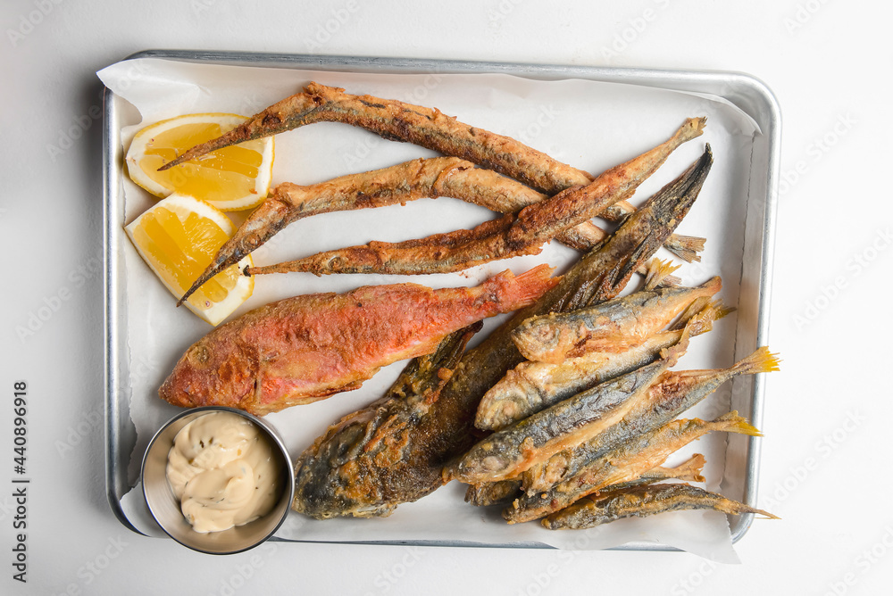 Fried fish with sauce and lemon served on a metal tray over white background.