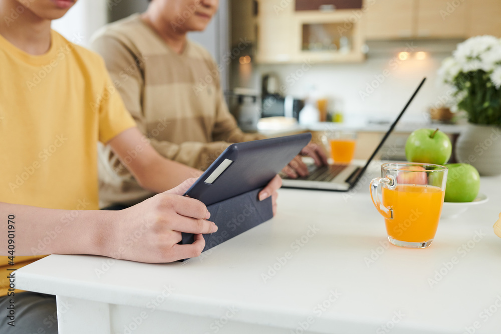 Teenage boy sitting at kitchen table next to his father working on laptop, drinking juice and watching show or educational video on digital tablet Teenage boy sitting at kitchen table next to his father working on laptop, drinking juice and watching show or educational video on digital tablet