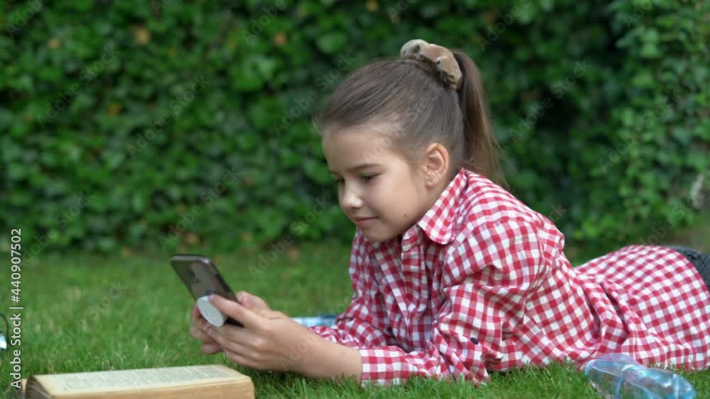 Little beautiful Caucasian lady lies on the green grass in the park and communicate with friends through the application on the smartphone. Little beautiful Caucasian lady lies on the green grass in the park and communicate with friends through the application on the smartphone.