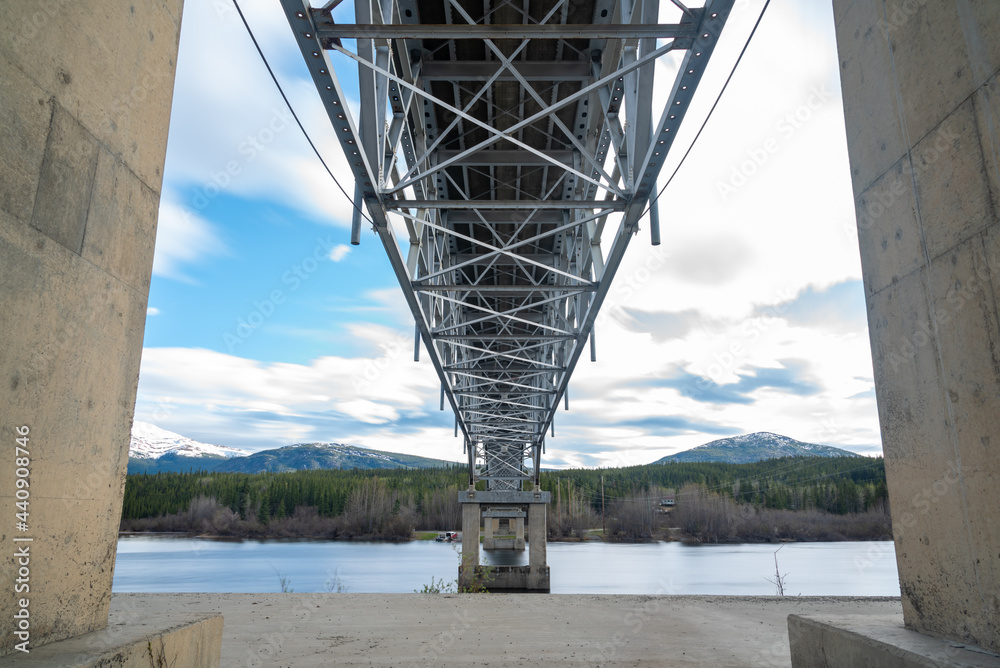 Below the magnificent Johnsons Crossing Bridge over Teslin River in northern Canada during spring, summer time with steel beams running above and blue sky in background. 