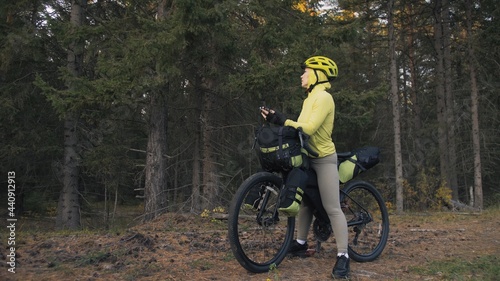 The woman travel on mixed terrain cycle touring with bike bikepacking outdoor. The traveler journey with bicycle bags. Sportswear in green black colors. Magic forest park. Make a selfie smartphone.