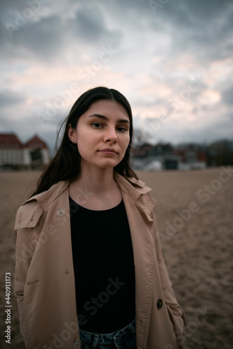 Trendy brown haired woman wears trench and jeans on the beach with beautiful classy looking hotel on the background. Vacation on the spring on the seaside concept.