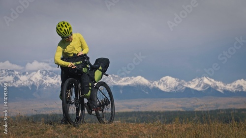 The woman travel on mixed terrain cycle touring with bikepacking. The traveler journey with bicycle bags. Sport tourism bikepacking, bike, sportswear in green black colors. Mountain snow capped.