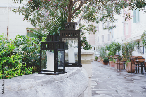 A flashlight candlestick, black vintage decorative lanterns with candles stands outdoor in city, in nature with flowers and olive tree. Home garden decoration, green lifestyle. 