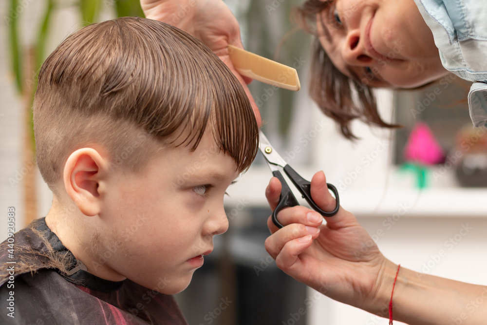a pretty young mother in a light blue dress with a comb and scissors in her hands cuts her son's hair at home. selective focus a pretty young mother in a light blue dress with a comb and scissors in her hands cuts her son's hair at home. selective focus