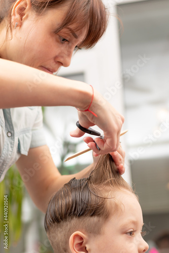 a pretty young mother in a light blue dress with a comb and scissors in her hands cuts her son's hair at home. selective focus