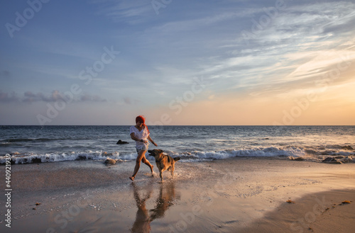 Happy red-haired woman in casual summer style running on the beach with her dog