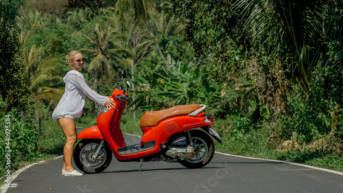 Woman on red scooter in white clothes drive on forest road trail. Dancing trip. One girl caucasian tourist in sunglasses dance, relax, rest near motorbike. Asia Thailand ride tourism. Motorcycle rent.