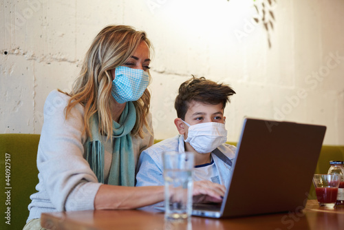 Mom and her son doing online education in a coffee shop, wearing masks during covid 19 pandemic