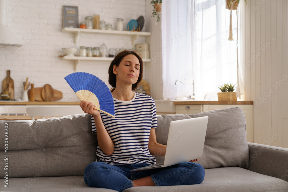 Tired millennial woman suffers from stuffiness and an inoperative air conditioner, waving blue fan sitting on couch at home working on laptop computer. Overheating high temperature, hot summer weather