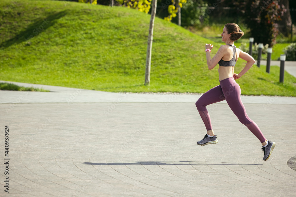 Young female runner, athlete is jogging in the city street in spring sunshine. Beautiful caucasian woman training, listening to music. Concept of sport, healthy lifestyle, movement, activity. Young female runner, athlete is jogging in the city street in spring sunshine. Beautiful caucasian woman training, listening to music. Concept of sport, healthy lifestyle, movement, activity.