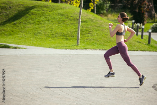 Young female runner, athlete is jogging in the city street in spring sunshine. Beautiful caucasian woman training, listening to music. Concept of sport, healthy lifestyle, movement, activity.