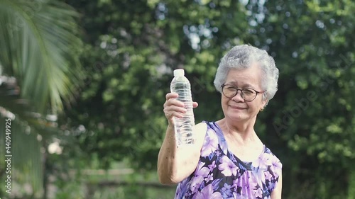 Beautiful senior woman with short gray hair holding a water bottle, smiling and looking at the camera while standing in a garden. Slow motion. Aged people and relaxation concept.