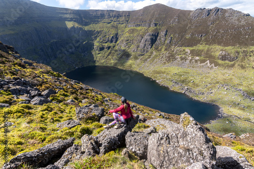 Woman wearing pink jacket sitting on stone looking at mountains and lake