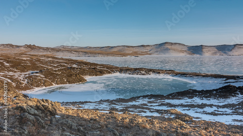 A small frozen heart-shaped lake. Near Baikal. There is snow on the surface of the ice. Around the land, devoid of vegetation. Mountain range against a clear sky. Siberia. Baikal region