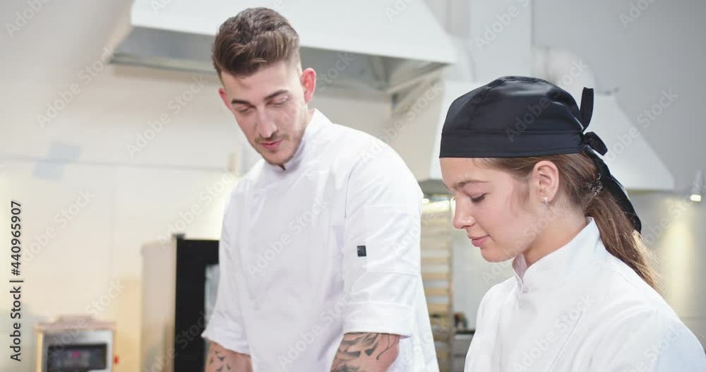 Close up. Side view of joyful handsome young man chef taching to prepare food meal at restaurant kitchen and speaking with female Caucasian cook colleague. Working process, culinary concept