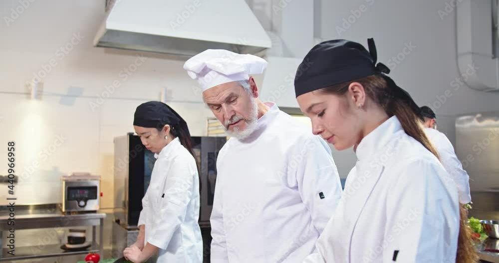 Close up of experienced famous senior Caucasian man chef preparing food at restaurant kitchen and speaking with female cook apprentice teaching to prepare meal. Working process, cookery concept Close up of experienced famous senior Caucasian man chef preparing food at restaurant kitchen and speaking with female cook apprentice teaching to prepare meal. Working process, cookery concept
