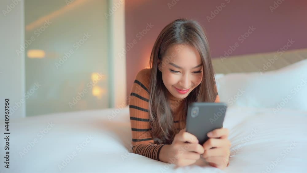 Asian woman holding phone in both hands and typing in messenger while lying on bed smiling Asian woman holding phone in both hands and typing in messenger while lying on bed smiling
