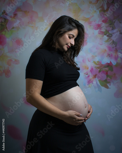 Pregnant young white woman looking at her belly of her first child a boy. Wearing a black outfit on a flowery background with both hands positioned around her belly.