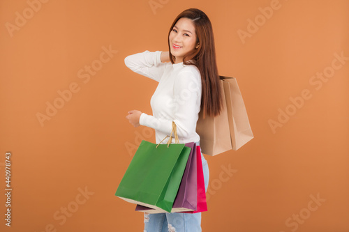 Beautiful young Asian woman wearing a white silk blouse, jeans, holding a shopping bag, smiling with a good mood, cheerful and cheerful in Holiday at the mall Center. With a brown background.