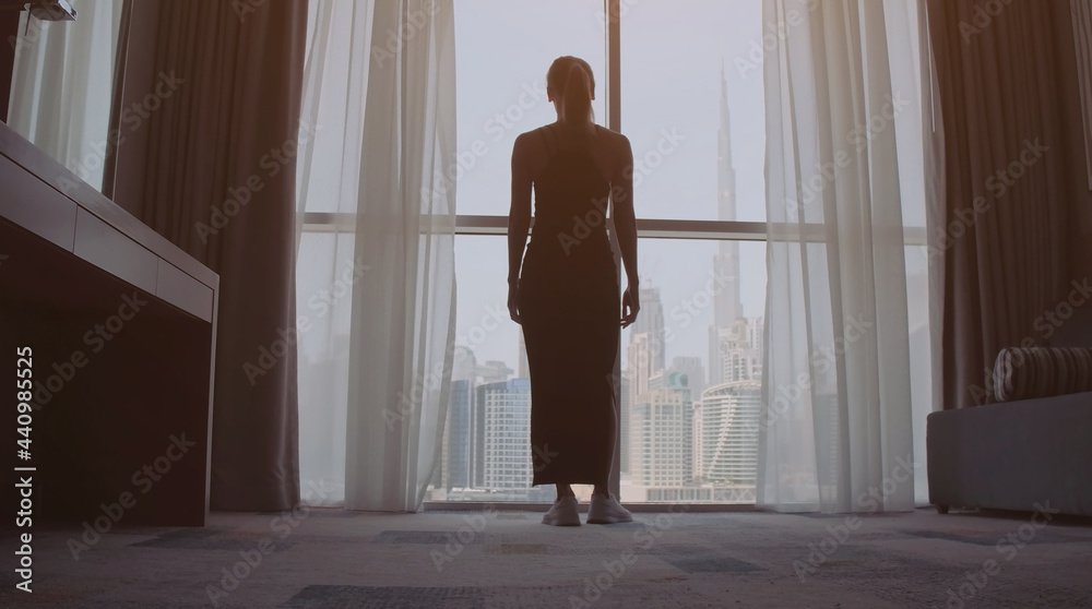 Back view of woman opening curtains and looking out of window to cityscape at hotel in the morning. Modern buildings at the Dubai city Back view of woman opening curtains and looking out of window to cityscape at hotel in the morning. Modern buildings at the Dubai city