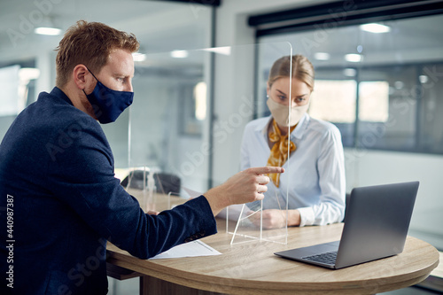 Business colleagues with face masks reading an e-mail on laptop while being separated with sneeze guard in the office.