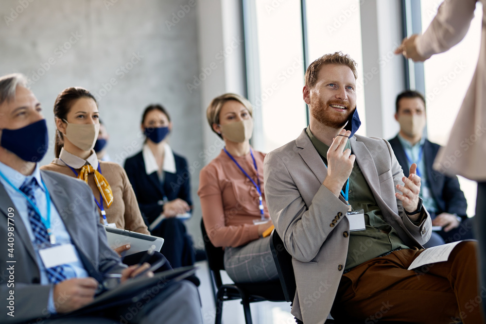 Happy businessman taking of protective face mask while talking to presenter during seminar. - obrazy, fototapety, plakaty 