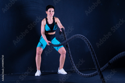 A brunette middle-aged woman in a tracksuit standing performs exercises for the arms, using the ropes. Indoor sports.