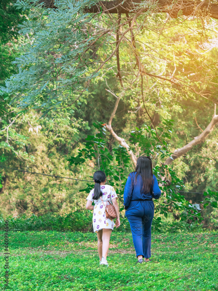 Back view of Asian mother and daughter walking through green garden. Mom and daughter walking together in park. Happy family spending time together outside in green nature. Enjoying nature outdoors. Back view of Asian mother and daughter walking through green garden. Mom and daughter walking together in park. Happy family spending time together outside in green nature. Enjoying nature outdoors.