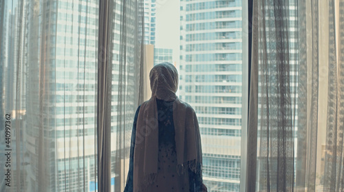 Silhouette of young woman opens curtains on the big window stretches arms and looking out her apartment on the city buildings during amazing sunrise. Dubai city