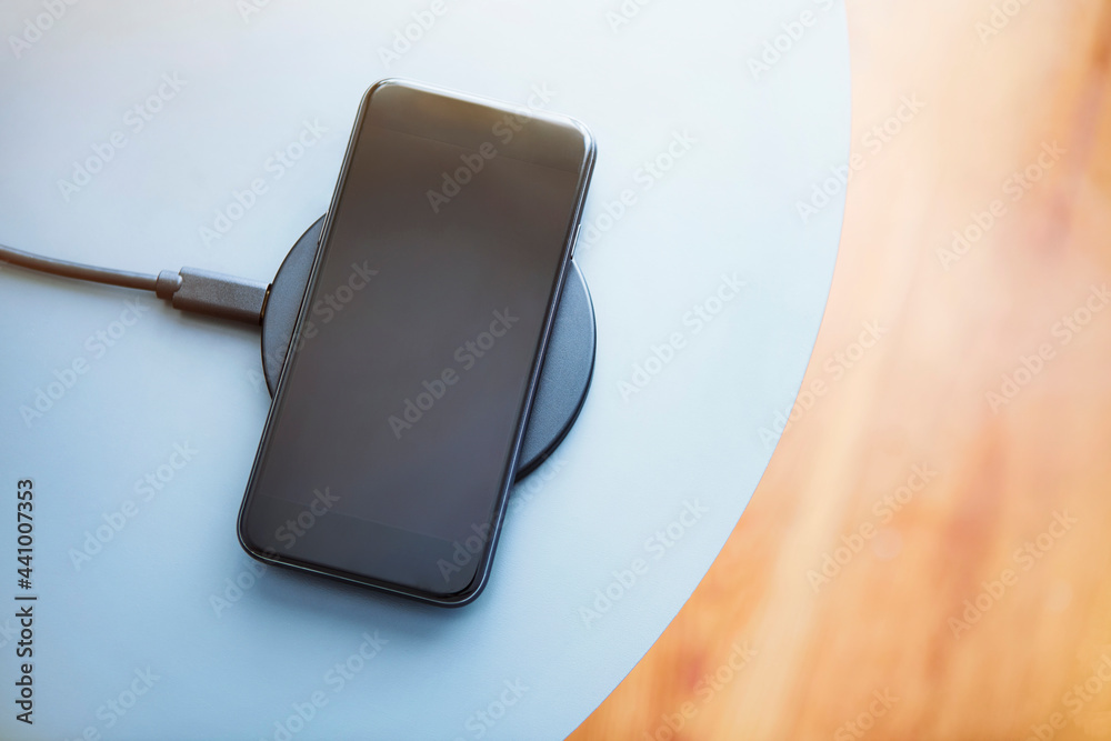 Top down view of smartphone lying on round charging pad. Light blue and wooden texture background. Top down view of smartphone lying on round charging pad. Light blue and wooden texture background.
