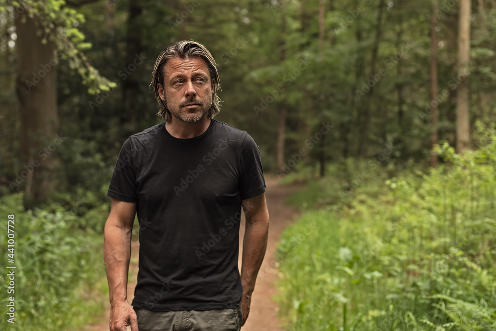 Blond man in a black t-shirt walks on a path in a lush forest.