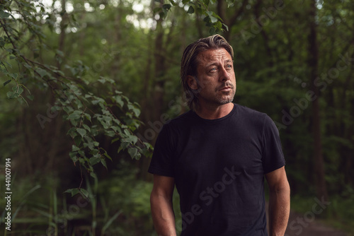 Blond man with stubble beard in a black t-shirt in a lush forest.