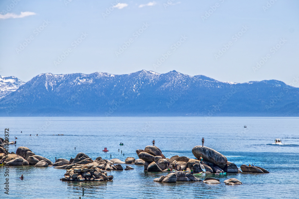 Tourists in water on paddleboards and climbing on rocks in blue Lake Tahoe with snow tipped mountains in distance