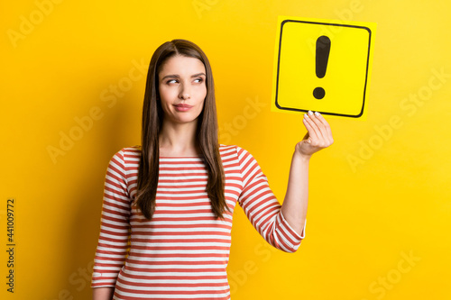 Portrait of attractive doubtful girl holding exclamation point card deciding isolated over bright yellow color background
