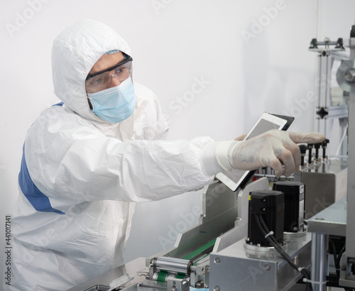 The engineer worker in protective suits, masks, and gloves is checking and controlling the machine in the manufacture of surgical masks for coronavirus. 