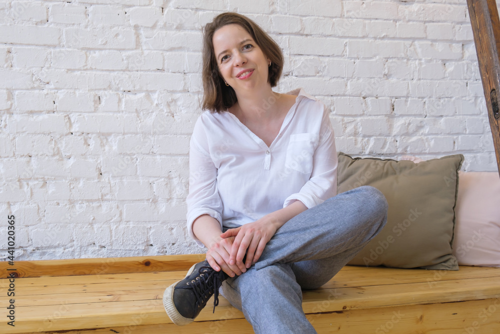Contented young woman in a loose white shirt, trousers and sneakers sits on a wooden bench in a loft on white brick wall background. Woman is in a relaxed pose and smiling. Contented young woman in a loose white shirt, trousers and sneakers sits on a wooden bench in a loft on white brick wall background. Woman is in a relaxed pose and smiling.