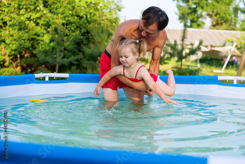 Father teaching his toddler daughter how to swim. Child swimming lesson. Cute little girl with father in summer metal frame pool at home in the yard. Pool time. Home vacations.  © Rodica