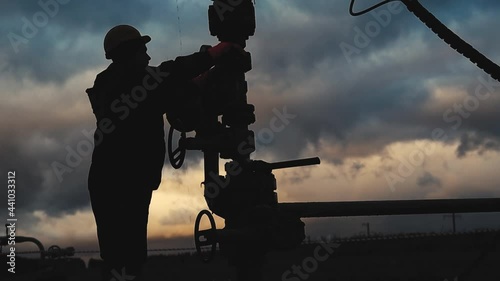 An employee in overalls and a helmet is carrying out repairs and maintenance of an oil well. Silhouette on the background of the evening sky