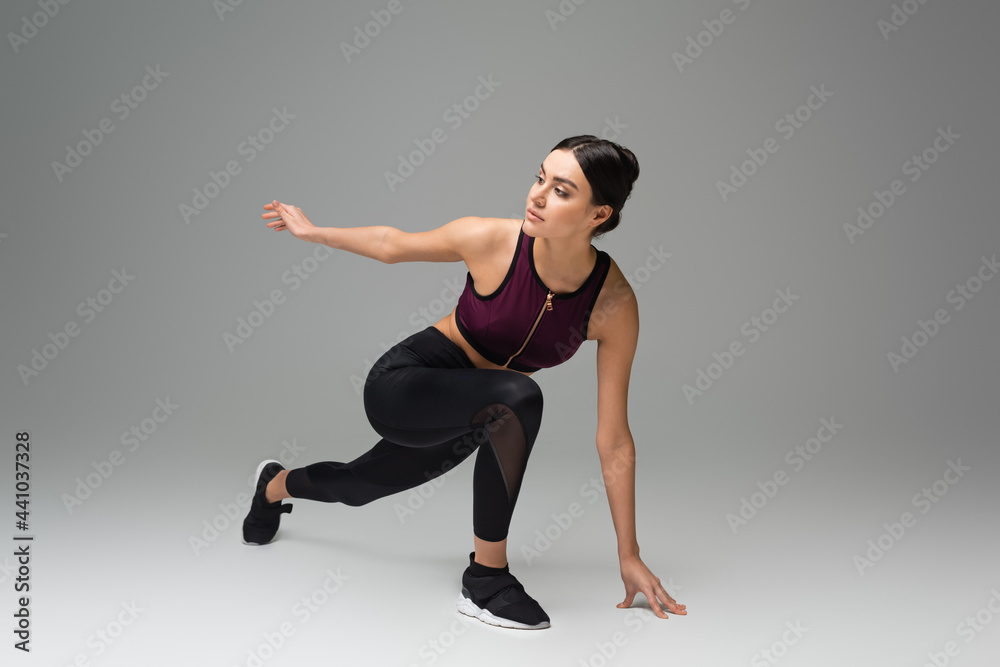 young sportswoman looking away while stretching on grey background.
